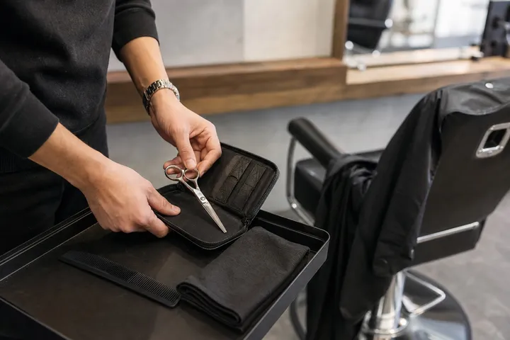 Hands lifting a closed shear from a protective case beside a salon chair.